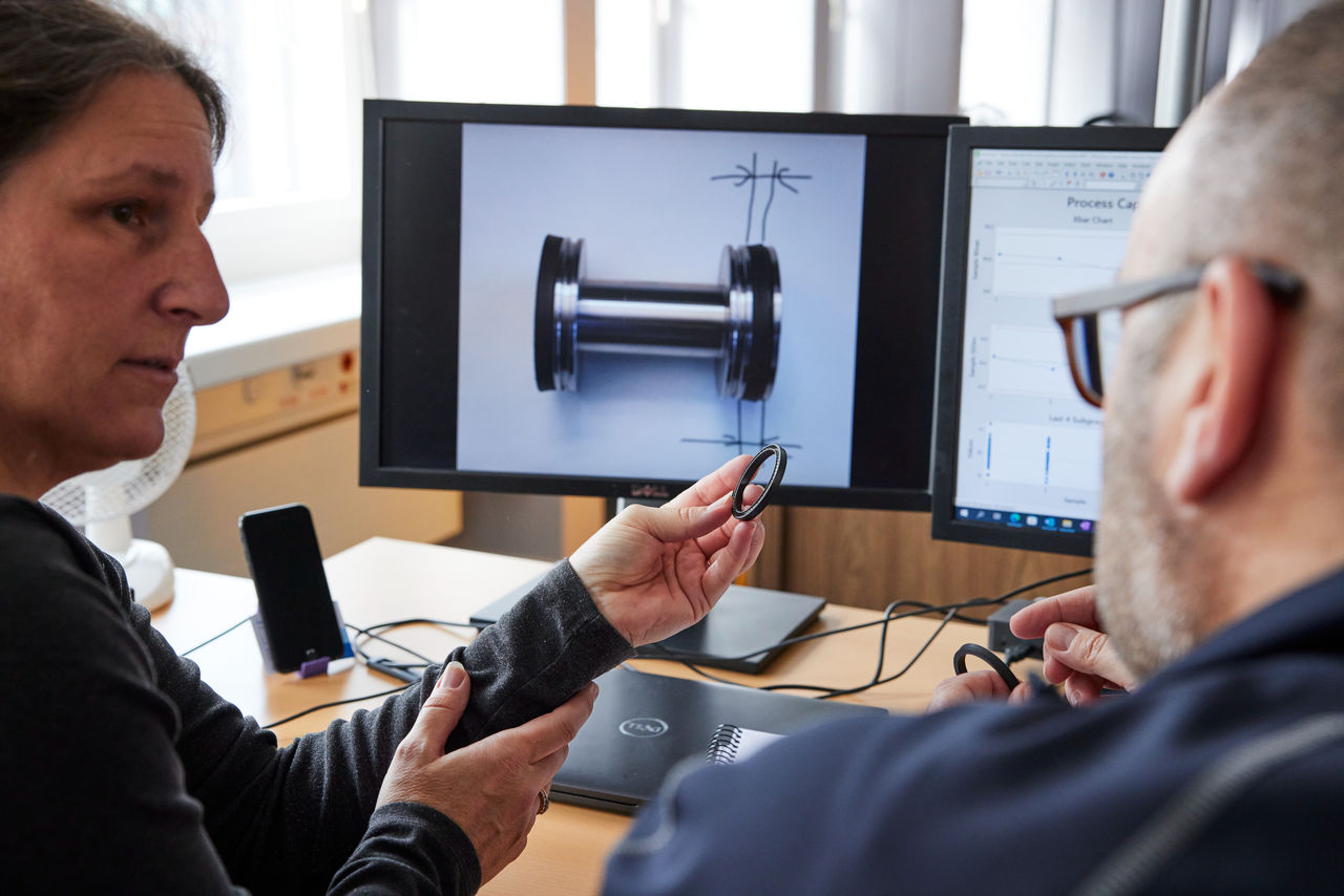 Two engineers examining a rubber seal in an office setting, with technical diagrams and a 3D image of a mechanical part displayed on dual computer monitors.