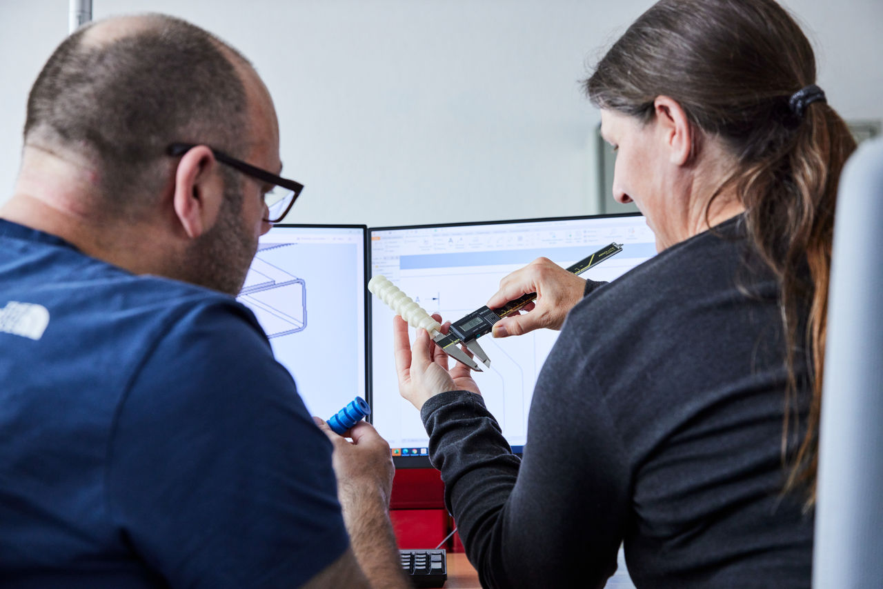 Two engineers reviewing a 3D-printed component, with one using digital calipers to take measurements while CAD drawings are displayed on dual monitors in the background.